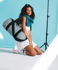 A woman kneels on the floor against a blue background, posing with a large grey Bagbase BG284 slung over her shoulder. She wears a white dress, a green jacket, and white sneakers with striped socks. Bright lights and a photography stand are visible on the sides.