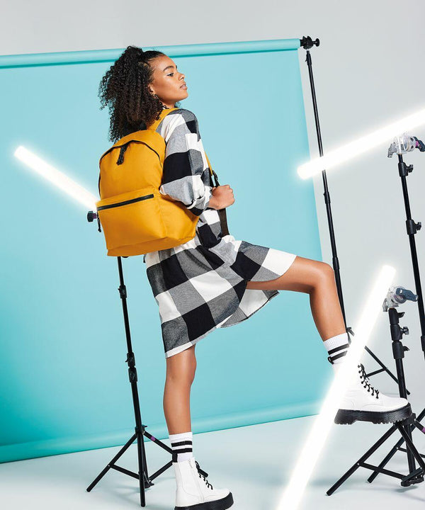 A person with curly hair wears a black and white checkered dress, white ankle boots, and a yellow Bagbase BG285. They step up onto a low platform in a brightly lit studio with a teal backdrop and vertical light tubes.