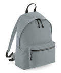 The BG285 backpack by Bagbase, shown on a white background, features a minimalist design in light gray with two zippered compartments. It also includes a front zipper pocket, black adjustable straps, and a top carry handle.