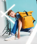 A person with curly hair wearing a blue shirt and jeans is posing on one knee against a turquoise background. They have a large yellow Bagbase BG286 backpack and white sneakers. Lighting equipment with fluorescent tubes shapes an "X" in the background.