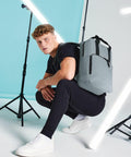 A man with short curly hair crouches in a studio setting. He is wearing a black outfit with a gray Bagbase BG287 backpack. There are lighting stands and white light tubes around him, and the backdrop is split with a blue and white color. He looks directly at the camera.