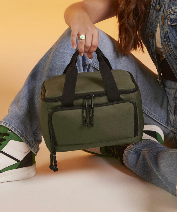 A person in casual attire, sitting with their knees bent, is holding a small olive-green Bagbase BG288 duffel bag with black zippers and handles. They are wearing a sleeveless denim jacket, light blue jeans, and white sneakers with green accents.