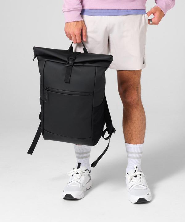 A person in a light purple sweatshirt, light shorts, white socks, and white trainers holds the Bagbase Athlux roll-top rucksack by its handle against a plain, light background.