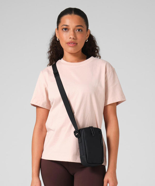 A woman with curly hair pulled back faces forward against a plain grey background, wearing a light pink T-shirt, brown trousers, and the Bagbase Athlux shoulder bag in black across her body.