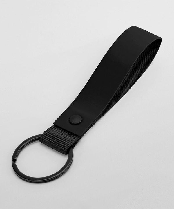 A black keychain features a wide loop made of sturdy, smooth material with a snap button near its base. Attached is a black metal key ring, suitable for holding multiple keys. The simple, sleek design is set against a plain white background. This product is the BG324 from Bagbase.
