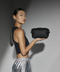 A woman with long hair in a sleek ponytail is standing sideways, holding a Bagbase BG330 with one hand. She is wearing a sleeveless black top and shiny silver pants. The background is plain and minimalistic.
