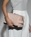 A person wearing a light gray short-sleeved shirt and high-waisted white pants is holding a large, rectangular, blush-colored Bagbase BG332. The bag features a black base. The background is a plain grayish-white wall.