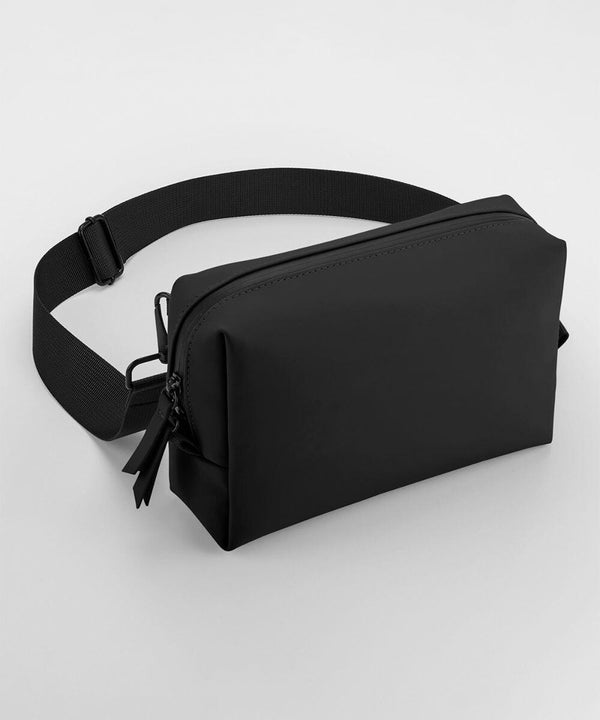 A Bagbase BG334 with two zipper pulls on the main compartment is shown. It has an adjustable strap and a minimalist design, lying on a flat white surface.