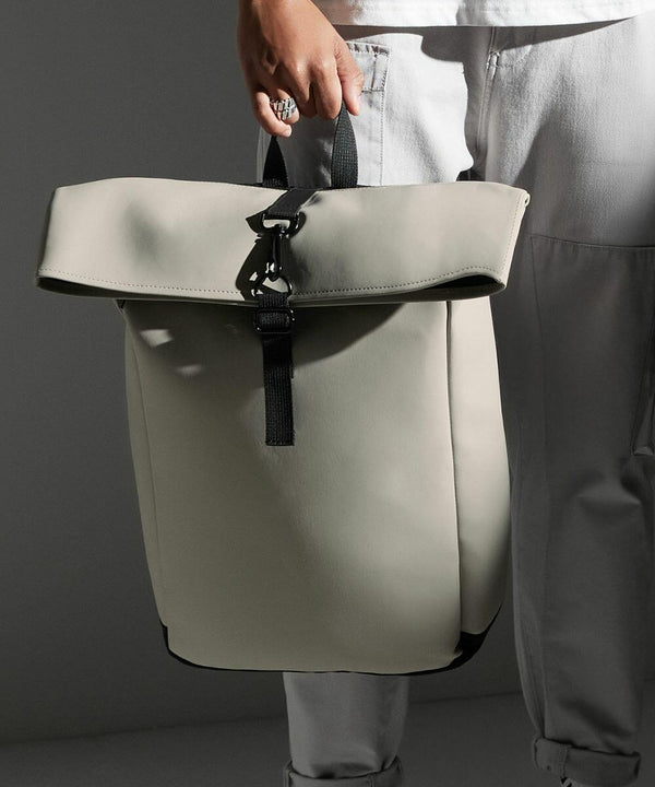 A person dressed in white holds a stylish, light gray Bagbase BG335 backpack by its handle. The Bagbase BG335 features a black strap with a metal buckle and has a clean, minimalist design. The background is a solid, dark gray.