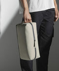 A person wearing a white shirt and dark pants is holding a Bagbase BG337 with a black zipper and small handle. The person's face is not visible in the image.