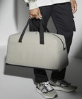 A person wearing a white shirt and black pants holds a beige Bagbase BG338 duffel bag with black handles. The individual also wears gray and white sneakers. The background is simple and unobtrusive, highlighting the bag.