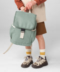 A child wearing brown shorts, a beige top with brown sleeves, striped yellow socks, and hiking shoes holds the Bagbase Kids adventure rucksack in light green with a black buckle. The background is plain light grey.