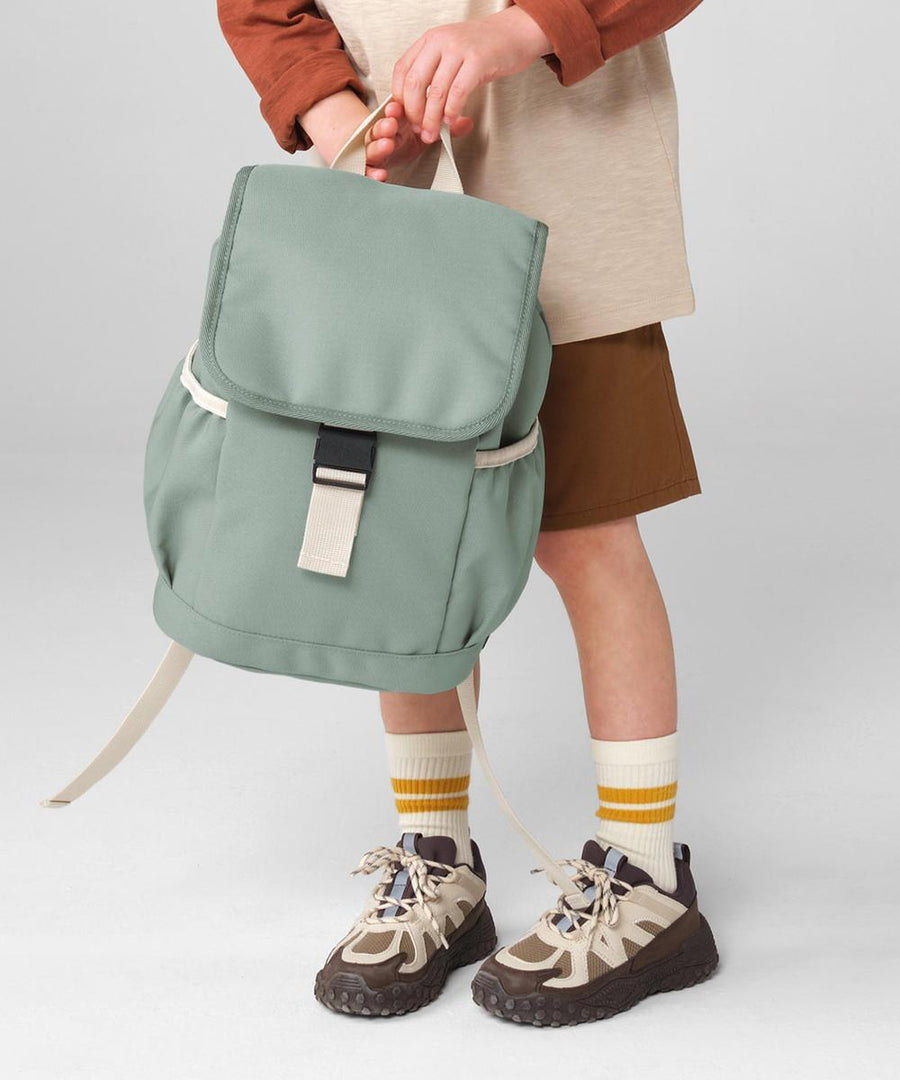 A child wearing brown shorts, a beige top with brown sleeves, striped yellow socks, and hiking shoes holds the Bagbase Kids adventure rucksack in light green with a black buckle. The background is plain light grey.