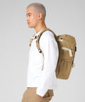 A person with short, fair hair is shown in profile wearing a white long-sleeve shirt, tan trousers, and the Bagbase Ramble rucksack against a plain light grey background.