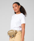 A woman with curly hair in a low ponytail wears a white T-shirt, beige trousers, and the Bagbase Ramble belt bag. She stands against a plain light grey background, looking at the camera with a neutral expression.