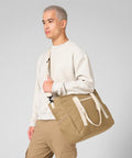 A person with short hair, wearing a light beige sweatshirt and tan trousers, carries the Bagbase Ramble tote—a large tan duffel bag with cream straps—over their shoulder against a plain light grey background.