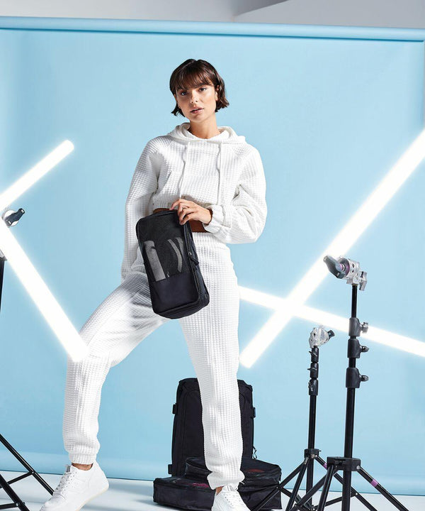 A person stands confidently in a white waffle-knit tracksuit, holding a Bagbase BG459. The backdrop is light blue, with white fluorescent light tubes arranged artistically around them. Photography equipment, including stands and lights, is visible.