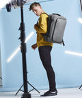 A person wearing a yellow sweatshirt, black trousers, and black trainers stands in a studio with a light blue backdrop, carrying the Bagbase Escape cabin backpack. Studio lights and equipment are visible in the foreground and background.