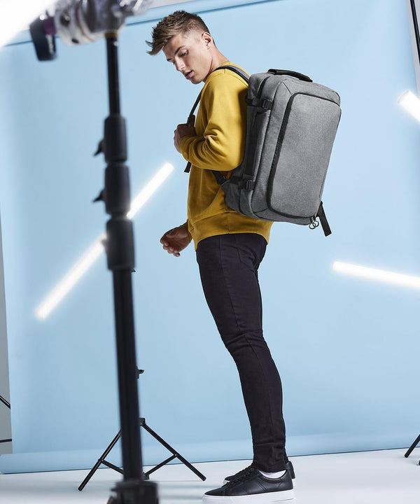 A person wearing a yellow sweatshirt, black trousers, and black trainers stands in a studio with a light blue backdrop, carrying the Bagbase Escape cabin backpack. Studio lights and equipment are visible in the foreground and background.