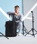 A young man in a grey tracksuit crouches beside a black Bagbase BG481 wheeled suitcase in a photography studio. He is surrounded by light stands with LED strip lights forming an X pattern in the background. He looks at the camera with a relaxed expression.