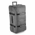 The BG483 by Bagbase is a sizable gray suitcase on wheels, featuring handles and external straps, shown standing upright against a simple white backdrop.