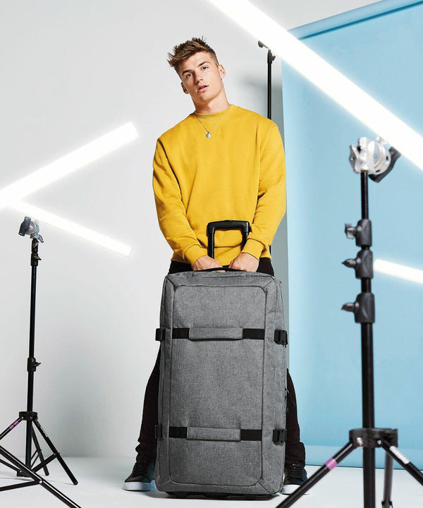 A person in a yellow sweatshirt stands behind a large gray Bagbase BG483 with black straps, holding its handle. The background features a blue panel to the right and vertical lights on stands.