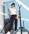 A stylish woman stands confidently in front of a blue backdrop, wearing a white sleeveless top, black sweatpants, and white sneakers. She holds colorful notebooks in one hand and rests her other hand on a large gray BG485 by Bagbase. Lighting equipment surrounds her.
