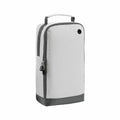 The Bagbase BG540 is a rectangular backpack in gray and white, featuring a top handle, a side zipper, and a ventilation hole. The bottom section of the backpack has a darker color, and it includes dual zipper pulls that have keychain rings.