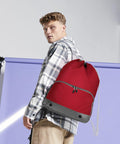 A person wearing a plaid shirt and khaki pants is carrying a red Bagbase BG542 backpack.