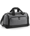 The Bagbase BG544 is a gray duffel bag featuring black accents and sturdy straps. It has a large main compartment secured with a zipper, two side pockets, and an additional front pocket with a zipper. An attached shoulder strap ensures easy carrying.