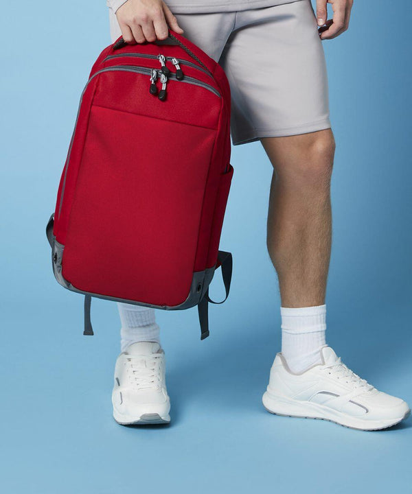 A person dressed in a light gray outfit holds a bright red Bagbase BG545 backpack in their hand. The individual is wearing white sneakers and white socks, standing against a light blue background. Only the lower torso and legs of the person are visible.