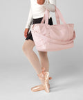 A person wearing a white long-sleeve top, black skirt, pink tights, and pink ballet pointe shoes stands on one foot holding a large light pink Bagbase Circuit gym bag. The head is not visible.