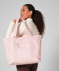 A woman with curly hair in a ponytail carries the Bagbase Circuit gym tote in light pink over her shoulder, styled with a beige long-sleeve top and brown leggings, standing against a plain light background.