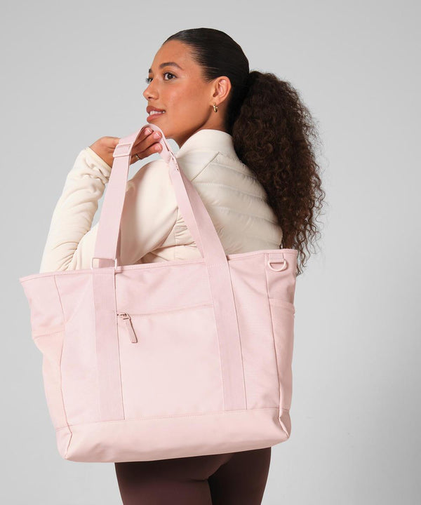 A woman with curly hair in a ponytail carries the Bagbase Circuit gym tote in light pink over her shoulder, styled with a beige long-sleeve top and brown leggings, standing against a plain light background.