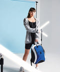A woman in a black dress and gray hoodie holds a blue and white Bagbase BG571 duffel bag. She stands in front of a blue backdrop with white light tubes around her.