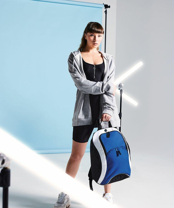 A woman in a black dress and gray hoodie holds a blue and white Bagbase BG571 duffel bag. She stands in front of a blue backdrop with white light tubes around her.
