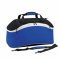 The Bagbase BG572 is a duffel bag in blue and black with white accents, equipped with handles and a detachable shoulder strap.