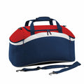 The Bagbase BG572 is displayed as a duffel bag in blue and red with a curved design, featuring white accents and navy straps. It includes a zippered main compartment, two top handles, a detachable shoulder strap, and small side pockets.