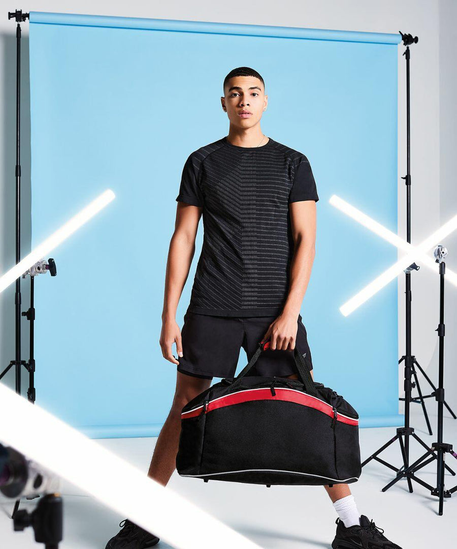 A person stands on a photoshoot set with a blue backdrop and lighting equipment, holding a black and red Bagbase BG572 sports bag, wearing a black shirt and shorts.
