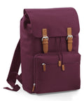 The Bagbase BG613 is a maroon backpack featuring a flap closure with two brown buckles, a front zippered pocket, and adjustable shoulder straps.
