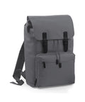 The BG613 by Bagbase is a gray backpack featuring a minimalist and functional design, with a flap top secured by two black buckles. It includes an external zippered pocket on the front and has adjustable shoulder straps for convenience.