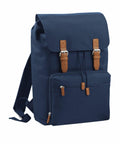 A dark blue canvas Bagbase BG613 backpack with brown leather straps and buckles, featuring a front zippered pocket and adjustable shoulder straps.