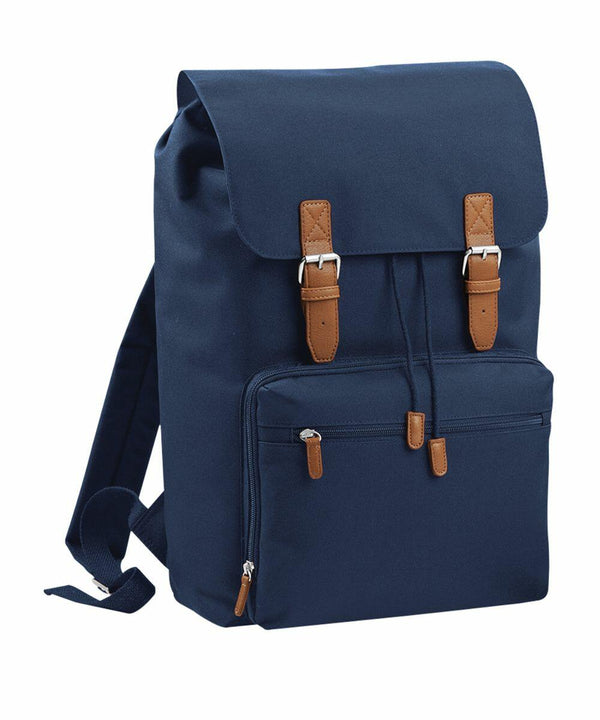 A dark blue canvas Bagbase BG613 backpack with brown leather straps and buckles, featuring a front zippered pocket and adjustable shoulder straps.