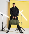 A person dressed in a black hoodie, black pants, and sneakers stands in a confident pose, holding a green Bagbase BG655. The background features a bright yellow wall with diagonal light beams and studio lighting equipment.