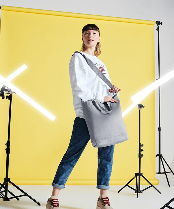 A person stands in front of a yellow backdrop holding a Bagbase BG673 tote bag. They wear a white long-sleeve shirt, blue jeans, and red sneakers. Studio lights are positioned on either side.