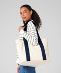 A woman with curly hair wears a white and black striped long-sleeve shirt, blue jeans, and carries the Bagbase Edit canvas tote with navy straps over her shoulder whilst looking towards the camera.