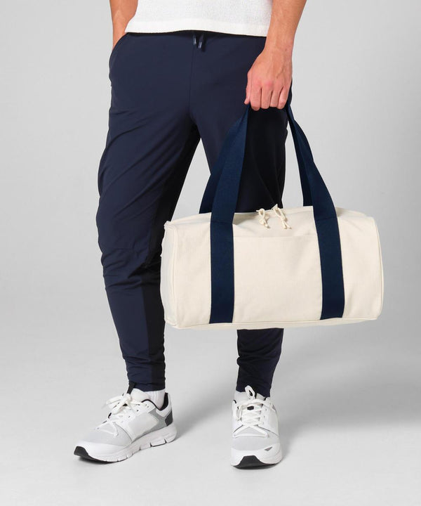 A person stands against a plain grey background holding the Bagbase Edit canvas duffle, a cylindrical cream-coloured bag with dark blue handles, while wearing dark blue joggers and white trainers.
