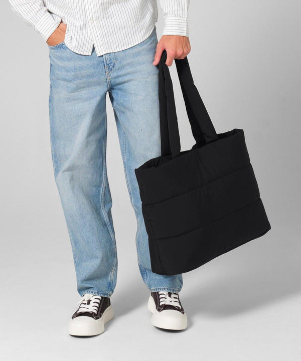 A person holds a large black Bagbase Quilted tote bag by its handles, styled with a white striped shirt, light blue jeans, and black-and-white trainers against a plain light background.
