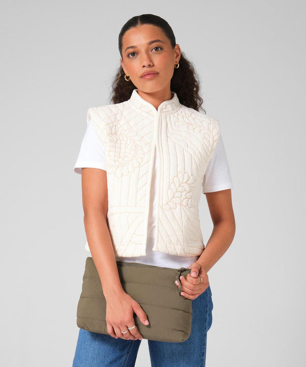 A woman with curly dark hair stands against a light grey background, holding the Bagbase Quilted Accessory Case in khaki, wearing a white quilted gilet over a white t-shirt and blue jeans.