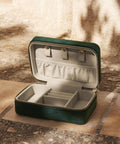 The Bagbase Velvet travel jewellery box, featuring a small green zipped design and light beige interior with multiple compartments and ring holders, is shown open on tiled stone flooring.